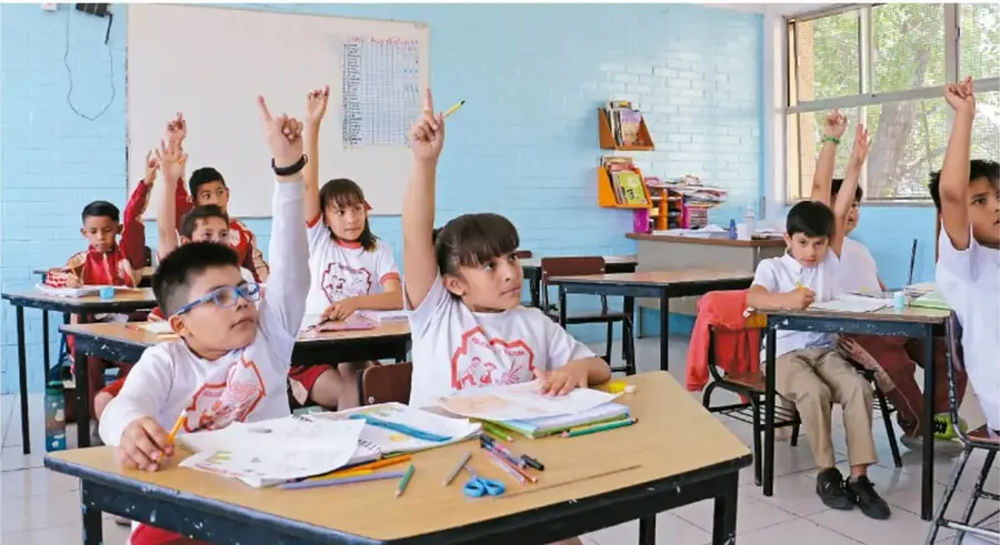 Fotografía de un salón de clases con estudiantes de primaria levantando la mano, sentados en sus pupitres, con cuadernos y colores sobre las mesas.