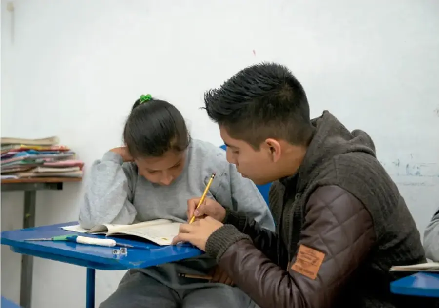 Dos estudiantes trabajando juntos en un pupitre azul, escribiendo en un cuaderno de forma colaborativa.