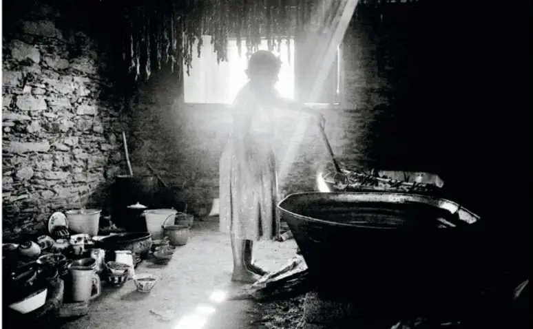 Fotografía en blanco y negro de un interior rústico de paredes de piedra; una mujer de vestido largo trabaja junto a pailas y recipientes, iluminada por la luz que entra por una ventana central.