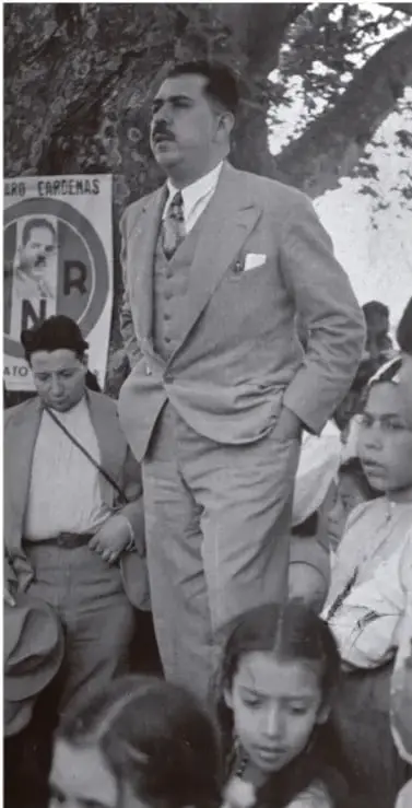 Fotografía en blanco y negro de Lázaro Cárdenas del Río de pie, con traje y corbata, acompañado de varias personas, entre ellas una mujer y niños. Al fondo se observa un cartel con la palabra 'CÁRDENAS'.