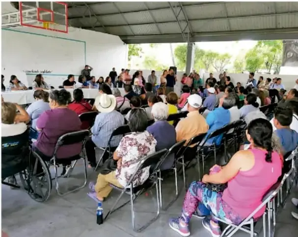 Fotografía de una asamblea o reunión comunitaria bajo un patio techado; hay muchas personas sentadas en sillas enfrentadas, de distintas edades, incluida una persona en silla de ruedas al frente a la izquierda; al fondo se ven árboles y paredes blancas.