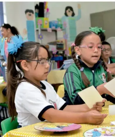 Dos niñas con listones azules y lentes sentadas en una mesa escolar durante la asamblea, con trabajos y adornos del salón al fondo.
