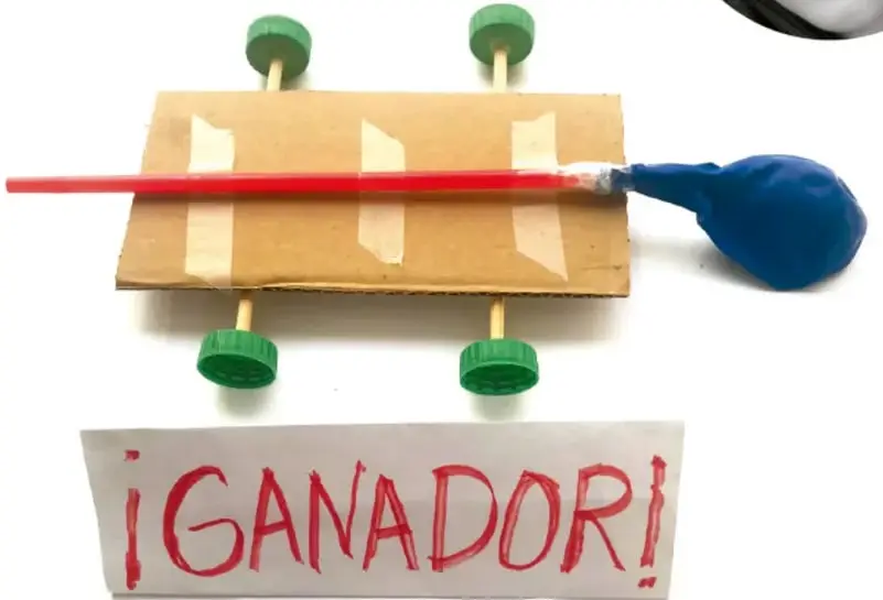 Carro de cartón con ruedas de tapas verdes y un globo azul sujeto a un tubo rojo, listo para propulsarse con aire.