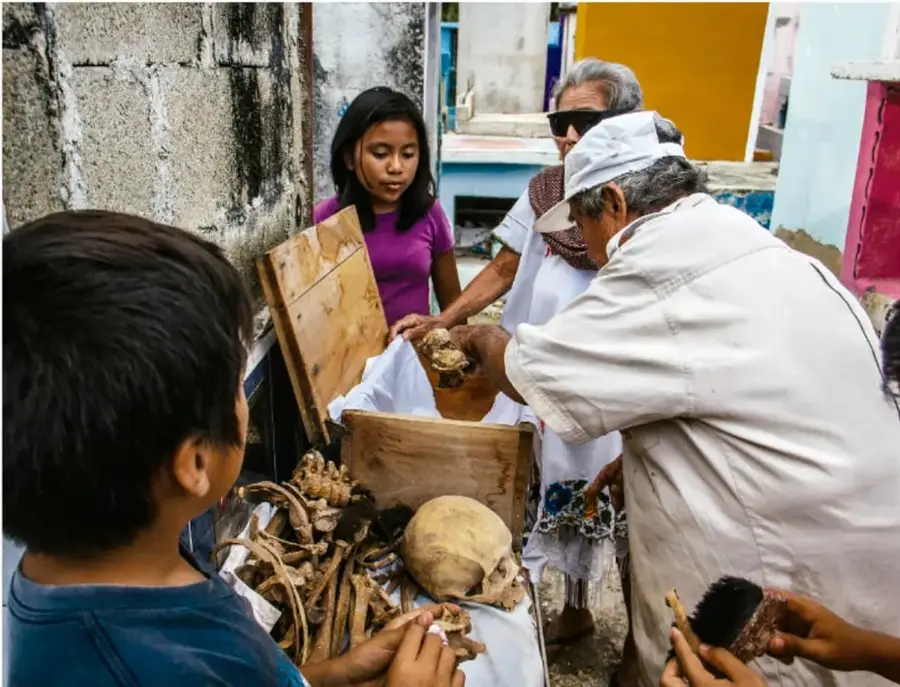 Fotografía de una familia en Pomuch limpiando los restos óseos de un familiar (incluye un cráneo y otros huesos) sobre un mantel blanco dentro de una caja de madera; participan niños, adultos y personas mayores, una de ellas con gorro blanco, en un patio al aire libre.