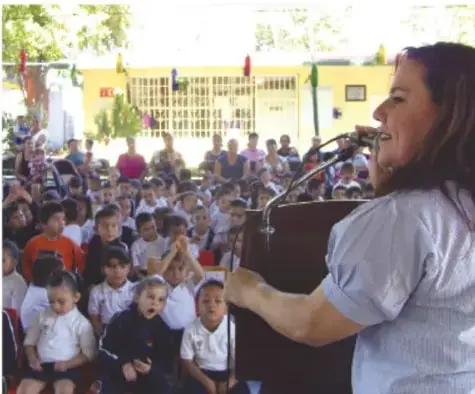 Fotografía de una mujer hablando frente a un micrófono ante una comunidad escolar reunida en el patio de una escuela.