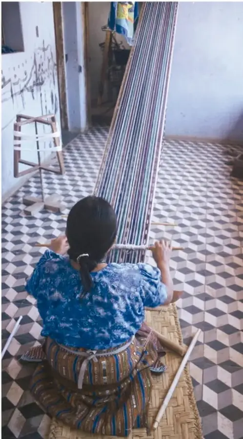 Mujer de espaldas tejiendo en un telar de marco dentro de una casa con piso de mosaico geométrico; viste blusa azul y falda tejida.