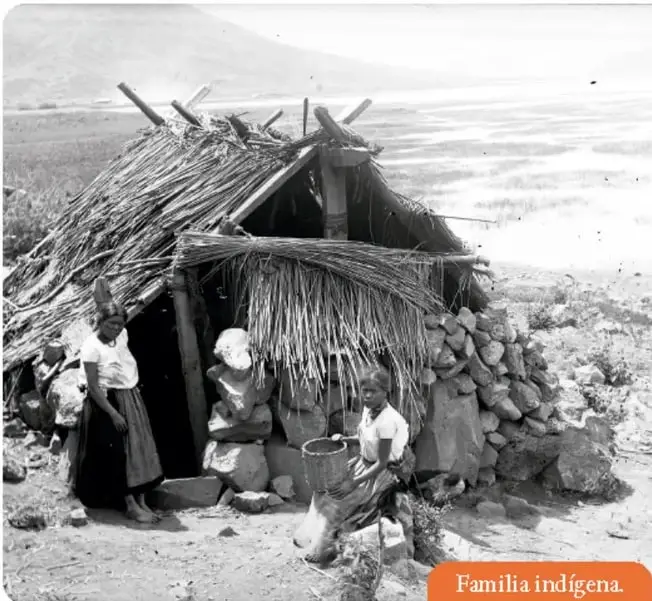 Fotografía en blanco y negro de una familia indígena frente a una choza de palma y piedra. Se observan tres personas: una mujer de pie, un niño sentado y una mujer joven con un canasto.