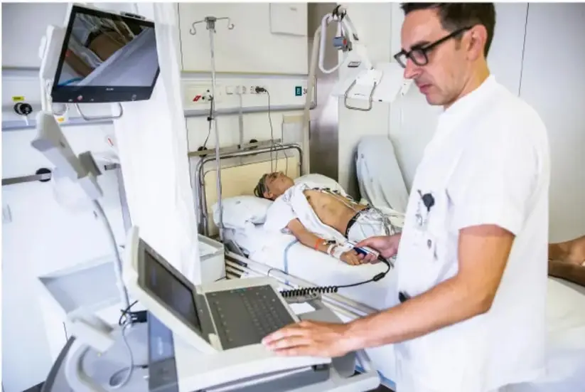 Fotografía de un profesional de la salud (doctor con cofia y lentes) operando una máquina de diagnóstico por imágenes en una habitación hospitalaria, mientras un paciente yace en la cama con electrodos conectados a un monitor.