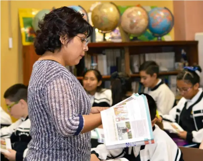 Fotografía de un aula de secundaria: una maestra de pie sostiene y muestra un cuaderno o tablero con imágenes a los estudiantes, quienes sentados en sus escritorios observan; al fondo hay globos terráqueos sobre una estantería.