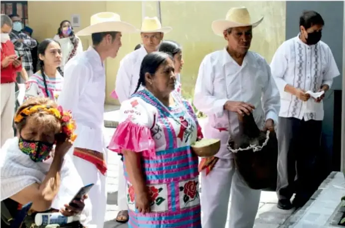 Fotografía de un grupo de integrantes de una comunidad indígena en una actividad comunitaria: personas con ropa tradicional bordada y sombreros de paja, algunas con cubrebocas; una mujer en el centro sostiene un recipiente metálico, un hombre a su derecha carga un cajón o canasta de madera. Ilustran la participación colectiva propia del tequio.