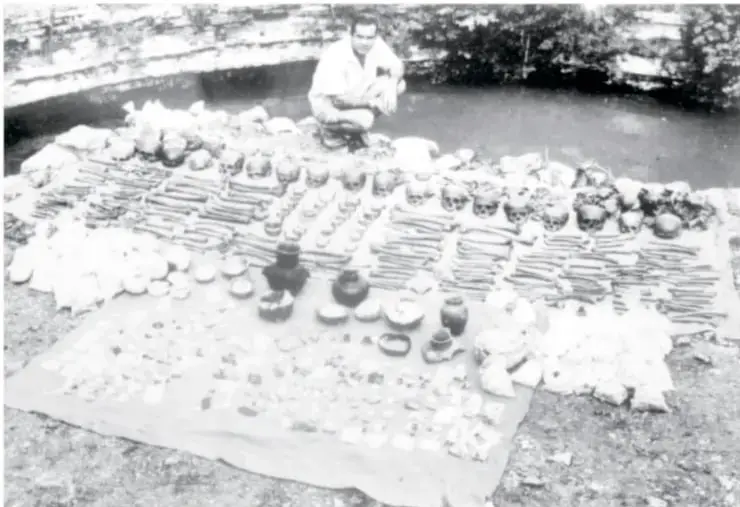 Fotografía en blanco y negro de una mesa o superficie con numerosas piezas arqueológicas pequeñas (vasijas, figuras y objetos) dispuestas ordenadamente; al fondo se observa una persona sentada junto a lo que parece ser un cenote o excavación.