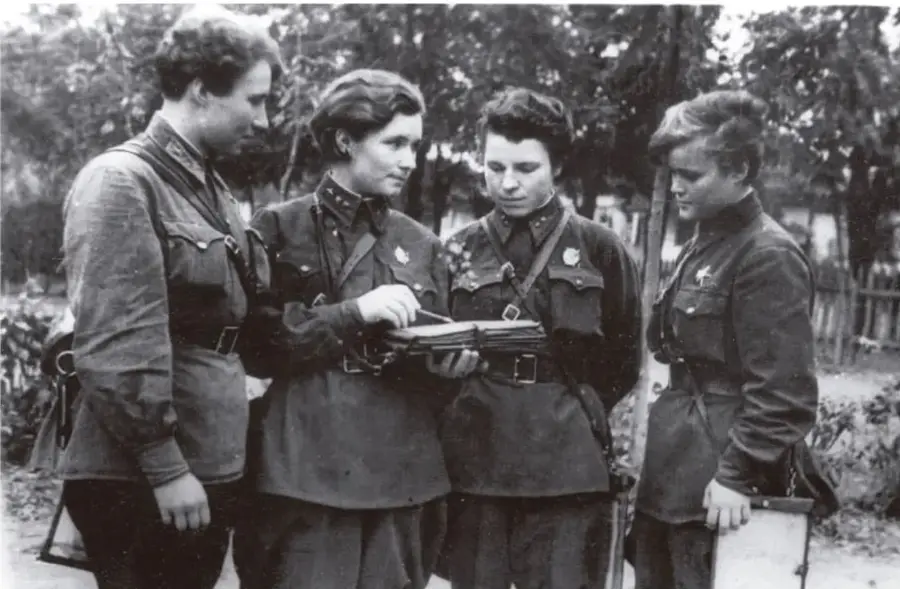 Fotografía en blanco y negro de cuatro mujeres con uniformes militares, reunidas alrededor de un mapa o documento que sostienen y observan.