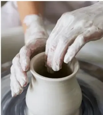 Manos de alfarero dando forma a una vasija sobre un torno, con los dedos cubiertos de barro blanco.