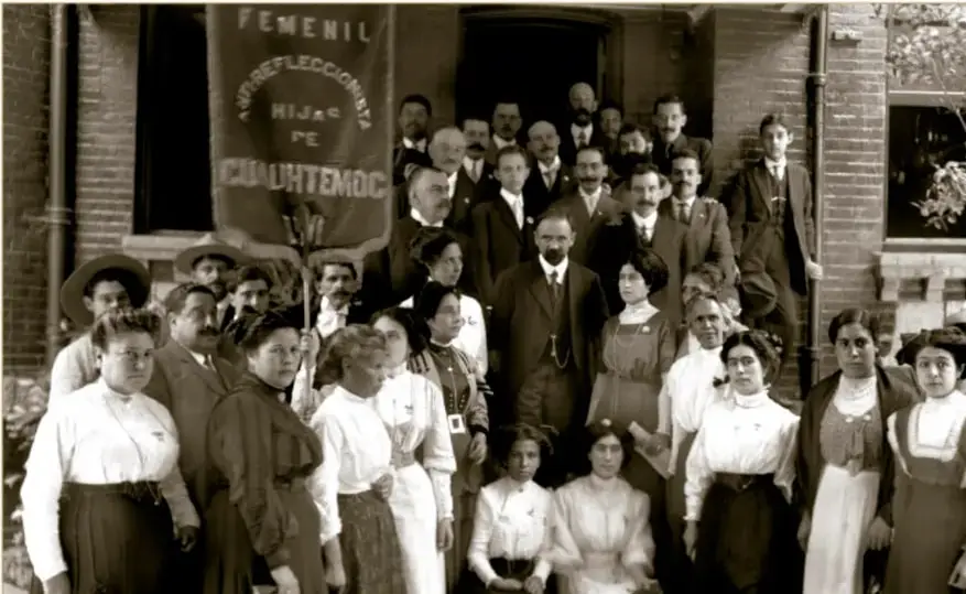 Fotografía histórica en sepia del Club femenil antirreeleccionista Hijas de Cuauhtémoc frente a la casa de Francisco I. Madero en México, el 2 de noviembre de 1911; se distingue un estandarte que dice 'FEMENIL ANTIRREELECCIONISTA HIJAS DE CUAUHTÉMOC' y un grupo numeroso de mujeres y hombres de la época.