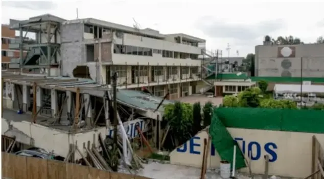 Fotografía de un edificio de tres niveles parcialmente colapsado, con escombros y una estructura de concreto dañada. Se observa una valla con la palabra 'JEFATURA' y vegetación alrededor.