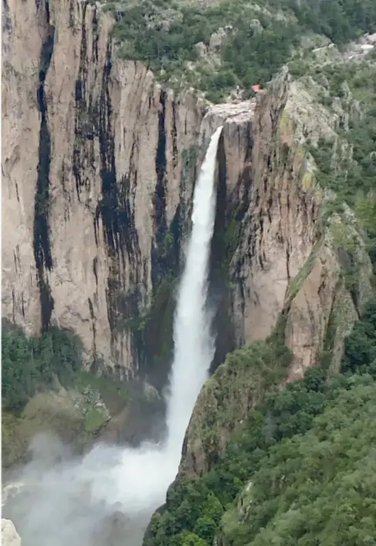 Fotografía vertical de una cascada alta y blanca que cae por paredes rocosas verticales entre vegetación verde; en la base se forma una nube de neblina.