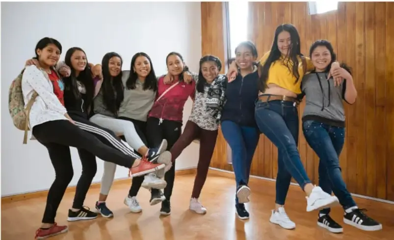 Fotografía de un grupo de nueve adolescentes (jóvenes estudiantes) posando juntas, sonriendo, con los brazos alrededor de unas de otras, en un espacio con piso de madera; representa la convivencia, la amistad y el sentido de pertenencia en la adolescencia.