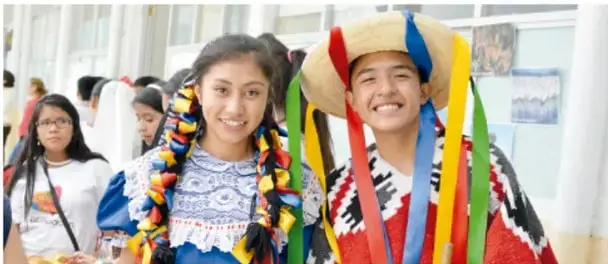 Dos jóvenes sonrientes disfrazados con trajes tradicionales coloridos (cintas, sombrero con listones) en un espacio escolar, con otros estudiantes al fondo.