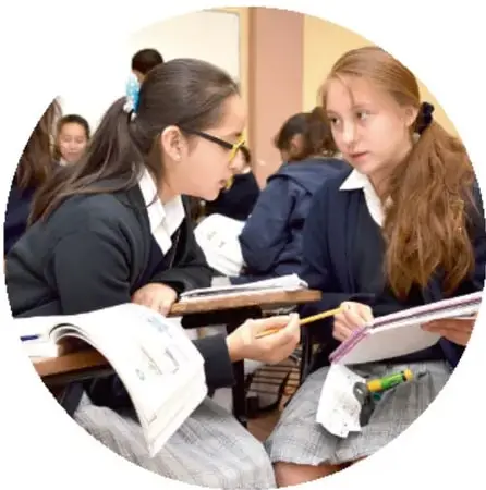 Dos estudiantes uniformadas trabajan juntas con cuadernos y lápices en el aula, conversando entre ellas.