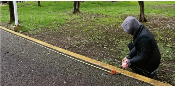 Persona arrodillada en un camino de parque midiendo con un flexómetro sobre una línea amarilla, junto a un poste blanco.