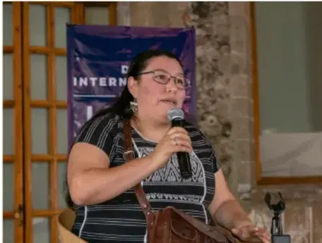 Fotografía de una mujer hablando con micrófono en un evento, probablemente una presentación o asamblea comunitaria; fondo con letrero parcial "INTERN".