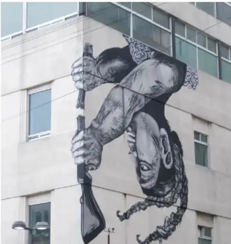 Mural en blanco y negro sobre un edificio: retrato realista de una persona con trenzas junto a una mano que sostiene un arma invertida, con una paloma en el hombro.