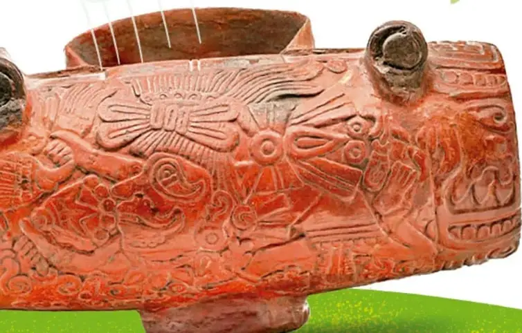 Vasija de barro rojo con figuras talladas de aves y rostros, con ranuras en la parte superior; parece un instrumento musical prehispánico.