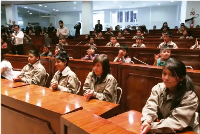 Grupo numeroso de estudiantes en un auditorio o aula amplia, sentados en filas, escuchando una actividad escolar.