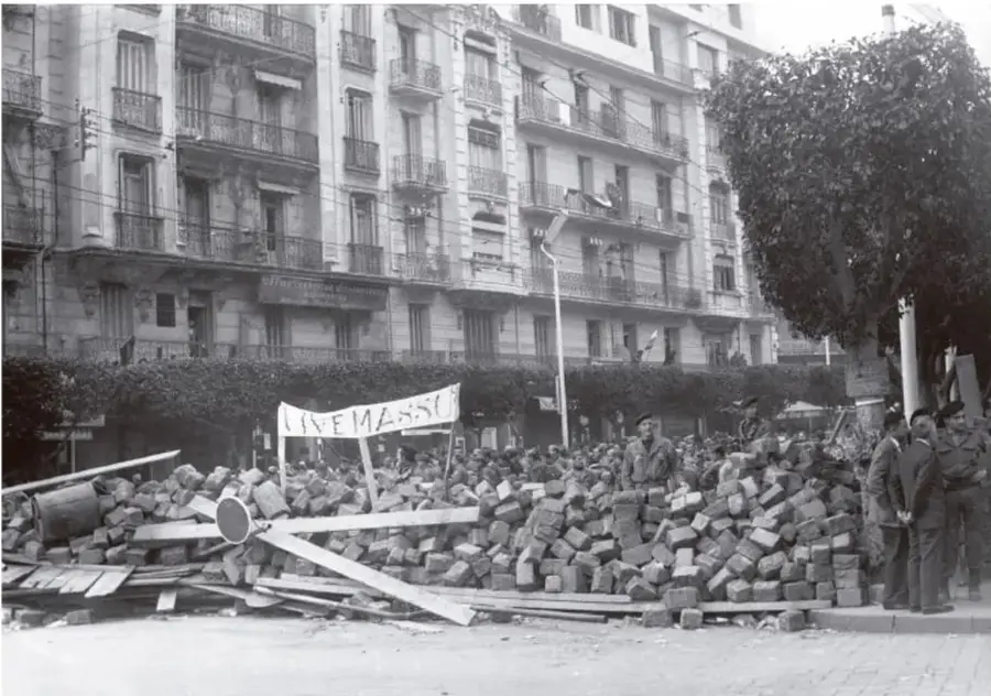 Fotografía en blanco y negro de una calle urbana con una barricada de escombros y ladrillos. Al fondo, edificios de varios pisos con balcones. En primer plano, una pancarta con la palabra 'LIVEMASSO' y varias personas, algunas uniformadas, observando.