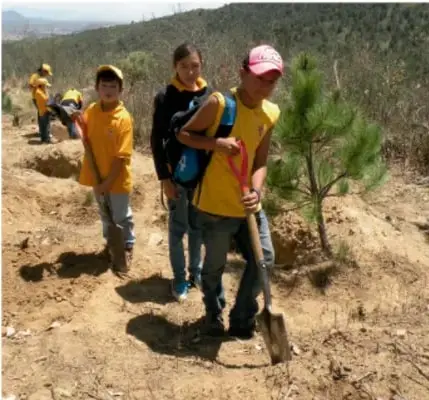 Fotografía de estudiantes con palas y cubetas en un terreno, acompañados de un adulto, observando el entorno.