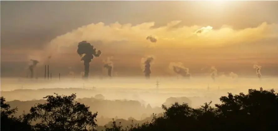 Fotografía al amanecer de chimeneas industriales emitiendo humo sobre un paisaje arbolado, representando la contaminación asociada al calentamiento global.