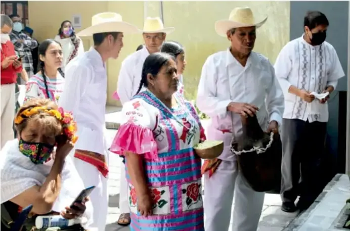 Fotografía de integrantes de una comunidad indígena, hombres con sombreros de paja y camisa blanca y mujeres con vestidos bordados de colores, participando en una actividad comunitaria en un espacio al aire libre.