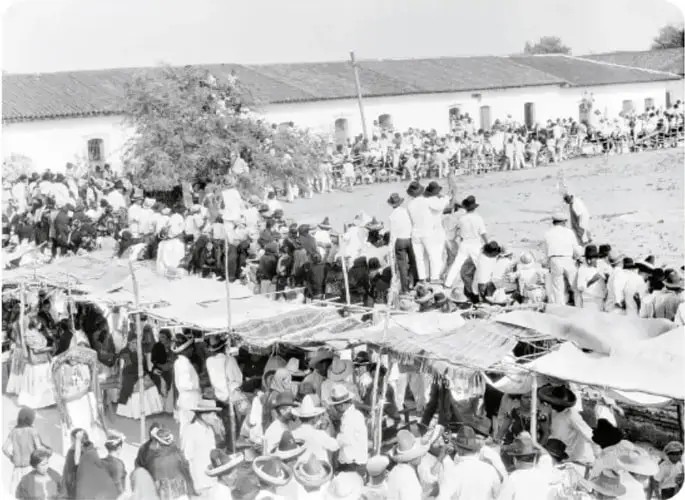 Fotografía en blanco y negro de una multitud reunida en un patio amplio: mujeres con trajes tradicionales (huipiles), hombres con sombreros, toldos de lona y edificios con techos de teja al fondo; pie: 'Fiesta en Juchitán, Oaxaca, ca. 1926'.