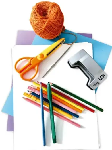 Fotografía de materiales escolares: un ovillo de hilo naranja, tijeras, una grapadora, lápices de colores, hojas de papel y cartulinas de colores sobre una superficie.