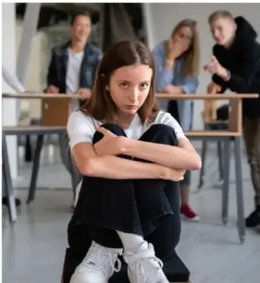 Fotografía de una adolescente sentada en el suelo con los brazos cruzados, con expresión de incomodidad; al fondo, tres compañeros la observan. Ilustra situaciones de vulnerabilidad o exclusión escolar.
