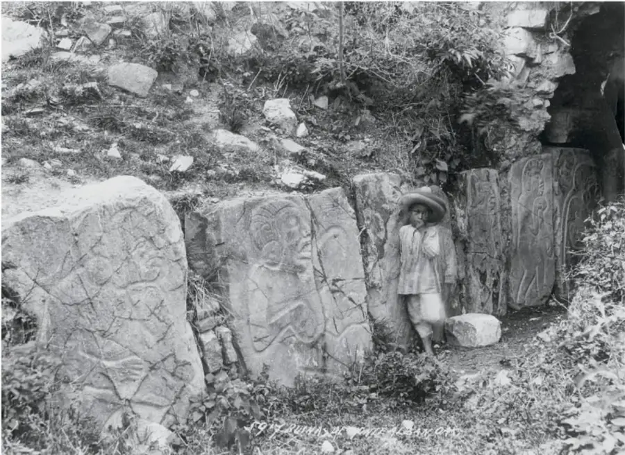 Fotografía antigua de una zona arqueológica: un niño con sombrero de paja se asoma entre grandes piedras talladas con grabados antiguos, rodeadas de vegetación. Lleva al pie una firma manuscrita ilegible.