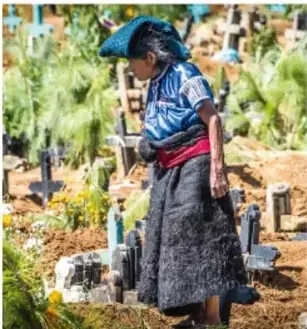 Mujer indígena con vestimenta tradicional y rebozo azul, de espaldas, en un panteón con flores, posiblemente durante una celebración conmemorativa (Día de Muertos).