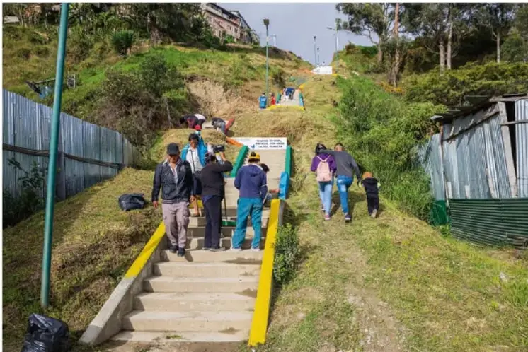 Fotografía de una comunidad en ladera con una escalinata de concreto amarilla; vecinos suben y bajan por las gradas, hay construcciones sencillas con láminas y vegetación, evidenciando un asentamiento popular.