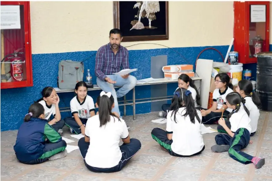 Fotografía de un docente sentado en una banca frente a un grupo de estudiantes de primaria que están en el suelo formando un círculo, en un patio escolar con muros azules y extintores rojos.