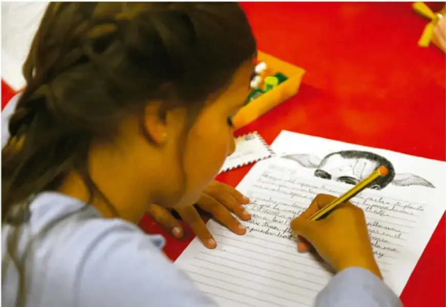 Fotografía de una niña de espaldas escribiendo una carta sobre una hoja rayada, con un lápiz, sobre una mesa roja. A su lado hay un sobre y una caja de colores.