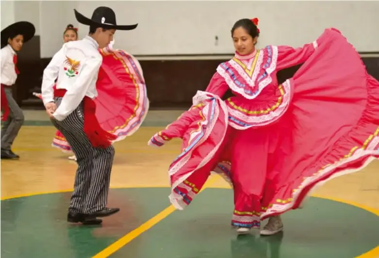 Fotografía de estudiantes bailando danza regional mexicana con trajes tradicionales (camisa blanca, pantalón de rayas con rebozo rojo y vestido amplio rosa) en el patio o gimnasio de una escuela.