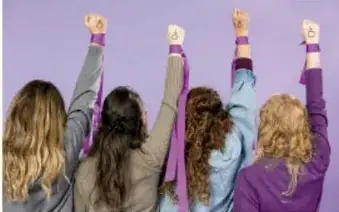 Fotografía de cuatro mujeres de espaldas con los puños en alto, envueltos con listones morados, símbolo del 8 de marzo y la lucha feminista, sobre fondo lila.