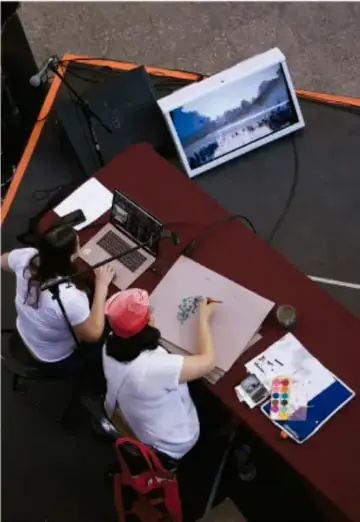 Fotografía de tres estudiantes trabajando alrededor de una mesa con una computadora portátil, una tablet que muestra una imagen de paisaje, papeles, colores y material de dibujo, preparando el material de su exposición.