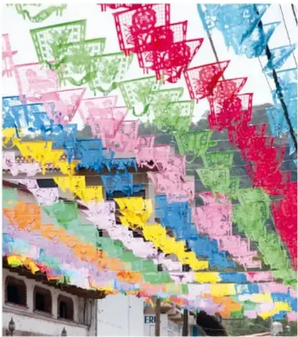 Muchas banderolas de papel picado de colores (rosa, verde, azul, amarillo) colgadas en calles con cielo nublado; foto de Carlos Hahn.
