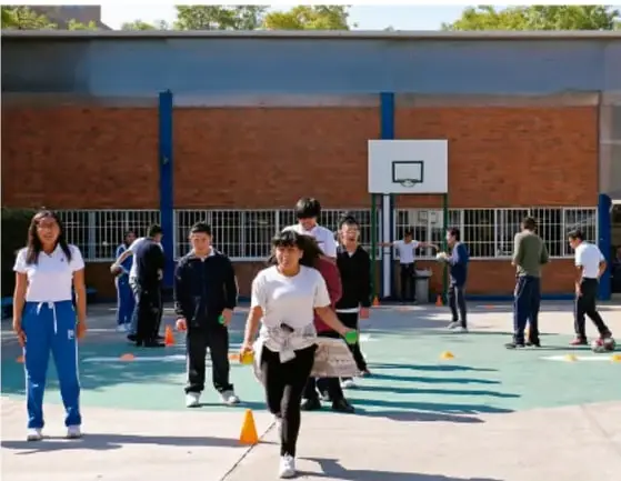 Fotografía de estudiantes en una cancha escolar al aire libre practicando una actividad física con conos naranjas; un grupo conversa al fondo junto a un aro de baloncesto.