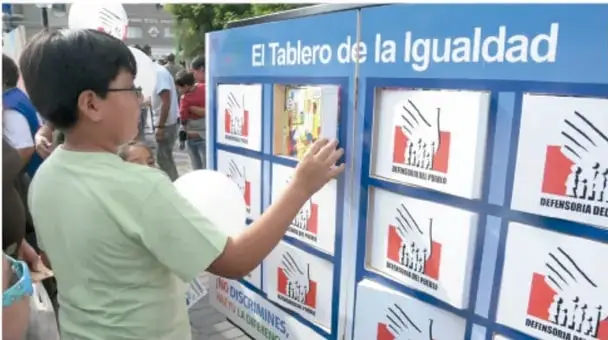 Niño de camiseta verde interactuando con "El Tablero de la Igualdad", tablero azul con tarjetas blancas de la Defensoría del Pueblo con imágenes de manos unidas.