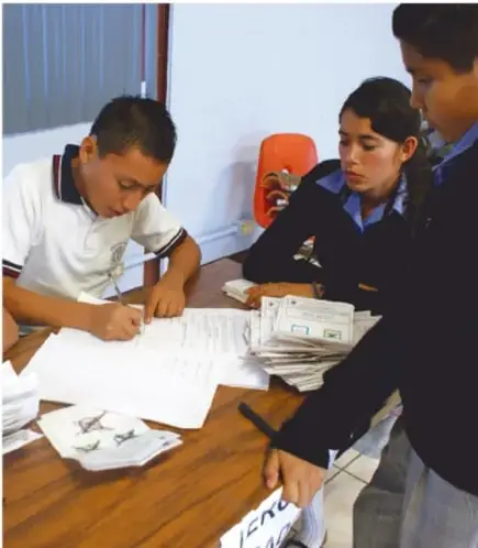 Jóvenes instalando o vigilando una urna electoral roja al aire libre, en una elección escolar o simulacro de votación.