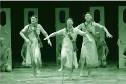 Fotografía a color (tono verdoso en la impresión) de la Compañía de danza Butoh Dairakudakan en Guanajuato: ocho bailarines con túnicas claras y banda cuadriculada; cinco arrodillados al fondo y tres de pie al frente sosteniéndose en un pie con los brazos extendidos; rostros de maquillaje blanco con párpados rojos, sobre un escenario de paneles verticales claros.