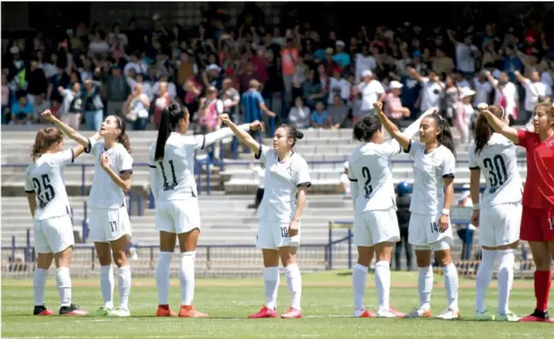 Fotografía de un equipo de fútbol femenil con uniforme blanco celebrando en el campo (jugadoras con los brazos en alto), portera con uniforme rojo y público en las gradas de fondo.