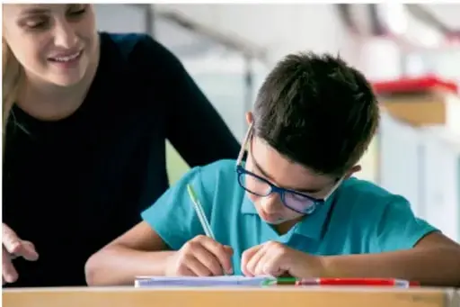 Niño con lentes escribiendo en una hoja, acompañado de una maestra.